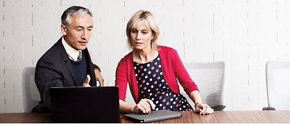 Een man en vrouw die aan een vergadertafel zitten en naar een laptop kijken.