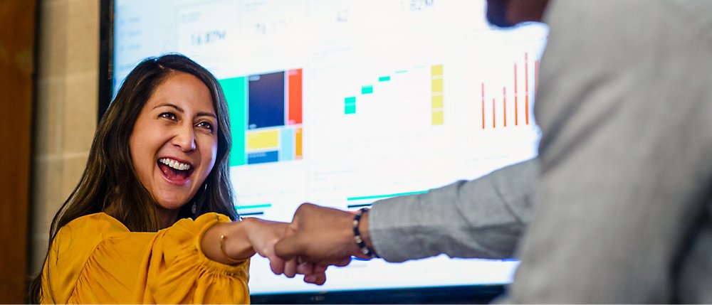 Eine Frau in einem gelben Oberteil lächelt und bietet einem Kollegen vor einem Bildschirm mit Grafiken und Diagrammen einen Fistbump.