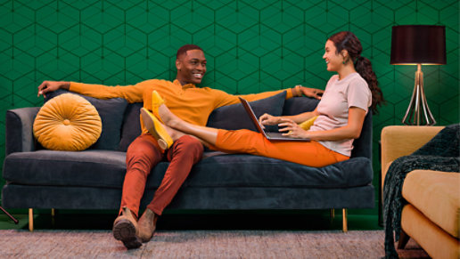 A couple  smiling and using a surface laptop while sitting on a couch in a colorful living room.