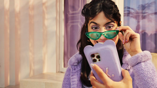A happy woman in green sun glasses and a purple sweater smiling and looking at a smartphone in a purple case.
