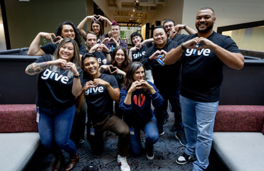A diverse group of Microsoft employees in a group photo wearing shirts that say, give and making heart signs with their hands.