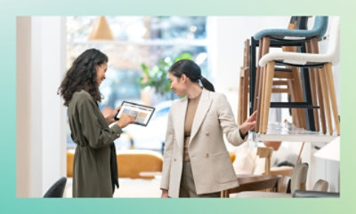 Two women in a furniture store with one holding a tablet and showing something to the other..