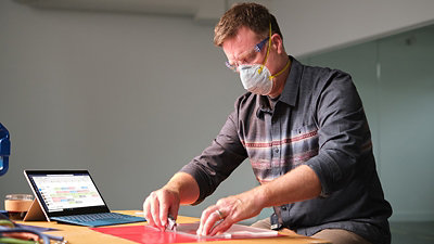 A person wearing P P E working at a desk with a tablet device to their right.