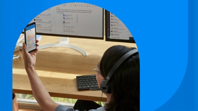 A person wearing a headset sitting at their desk and holding up a mobile phone.
