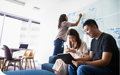 Drei Personen in einer Besprechung, von denen zwei auf ein Tablet schauen und die dritte auf einem Whiteboard schreibt.