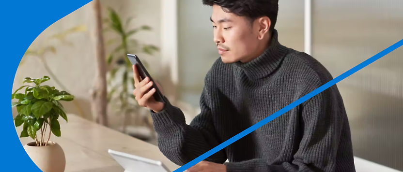 Hombre con suéter de cuello vuelto usando un smartphone en un escritorio con un documento y una maceta cerca.