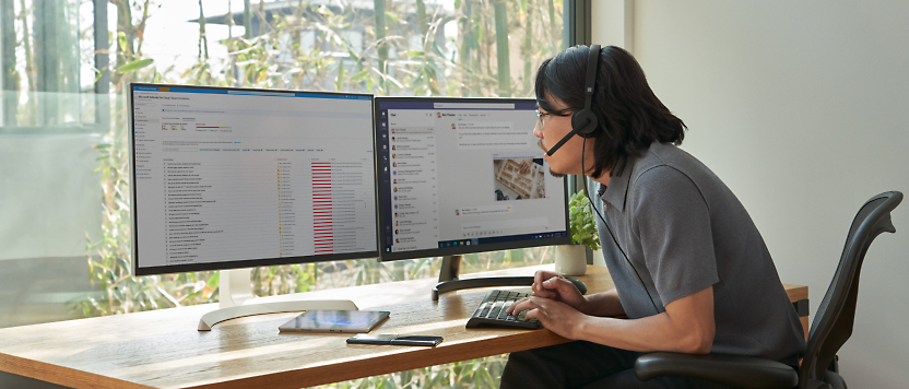 Een persoon met een headset werkt aan een bureau met twee beeldschermen die grafieken en e-mailberichten weergeven,