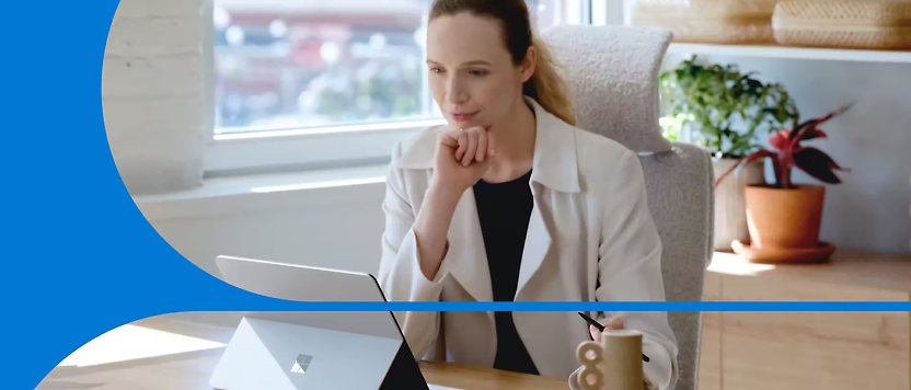Mujer con una chaqueta mirando atentamente a un portátil en una oficina con una planta y ventanas al fondo.