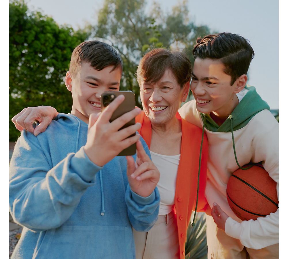 Two teenagers and an elderly woman smile as they look at a smartphone together outdoors.