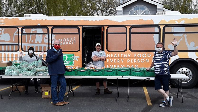 Four people stand behind a table with fresh produce in front of a mobile market bus that reads 'Healthy food, every person, every day.'