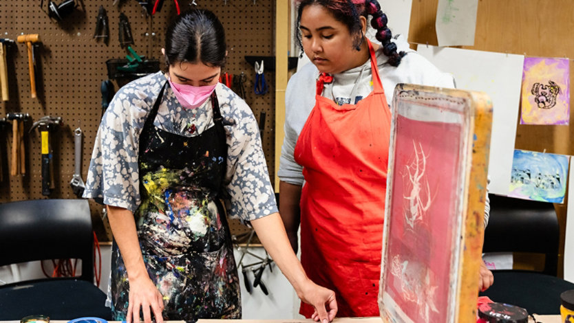 Two people in an art studio work with screen printing, surrounded by inks and screens. One explains while the other observes.