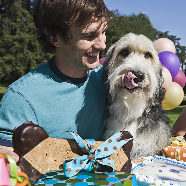 A man smiling with a dog at an outdoor celebration, with gifts and balloons in the background.