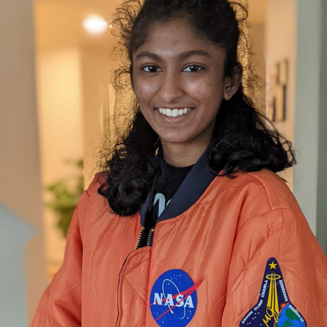 A smiling girl wearing an orange NASA jacket indoors.