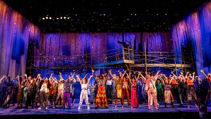 A large group of performers stands on a theater stage, arms raised, under colorful lights and falling confetti. The backdrop is deep blue.