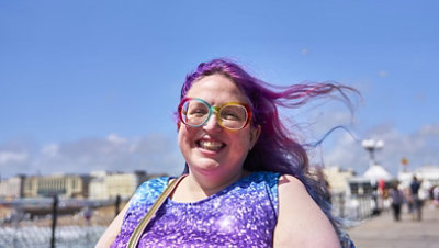 A smiling person with purple hair and colorful glasses enjoys a sunny day outdoors.