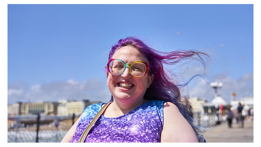 A smiling person with purple hair and colorful glasses enjoys a sunny day outdoors.