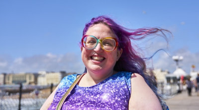 A woman with colorful glasses and purple hair outdoors on a sunny day.