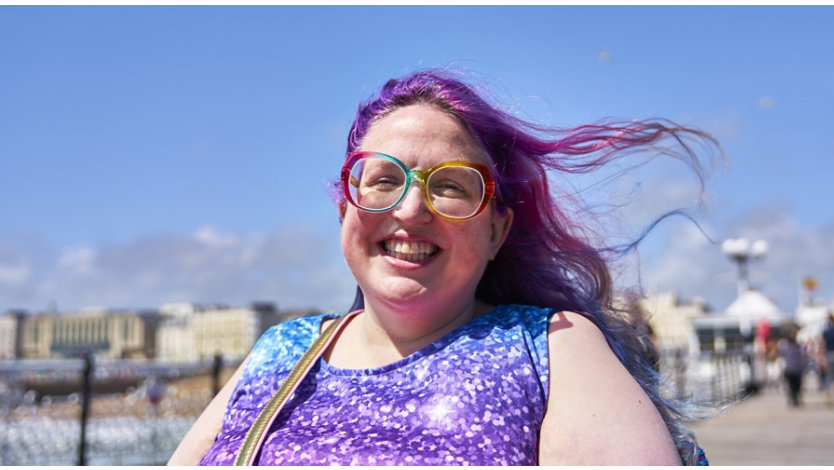 A woman with colorful glasses and purple hair outdoors on a sunny day.