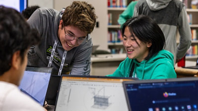 Students working on laptops in a library with bookshelves in the background.