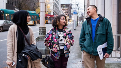 Three people walking down a city sidewalk engaging with each other, walking and talking. Two female and one male.