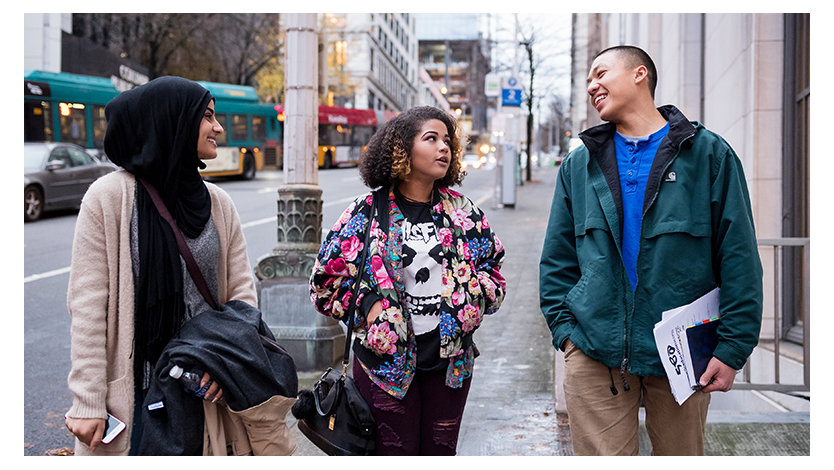 Three people walking down a city sidewalk engaging with each other, walking and talking. Two female and one male.