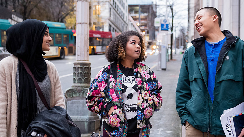 Group of three individuals walking down a city street talking and looking at each other.