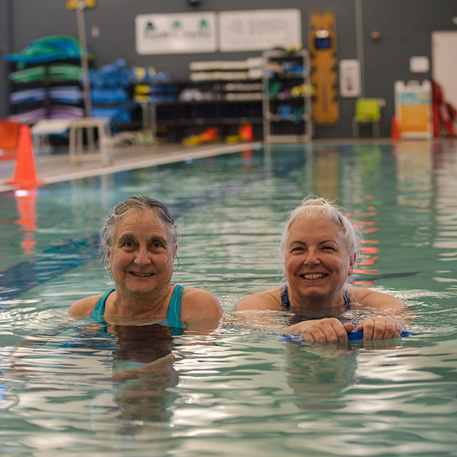 Two older women swimming in a pool together smiling.