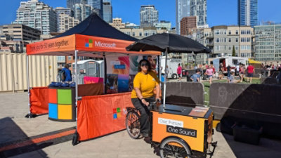 A woman with a tricycle cart at an outdoor event, with city buildings in the background.