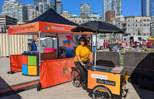 A woman with a tricycle cart at an outdoor event, with city buildings in the background.