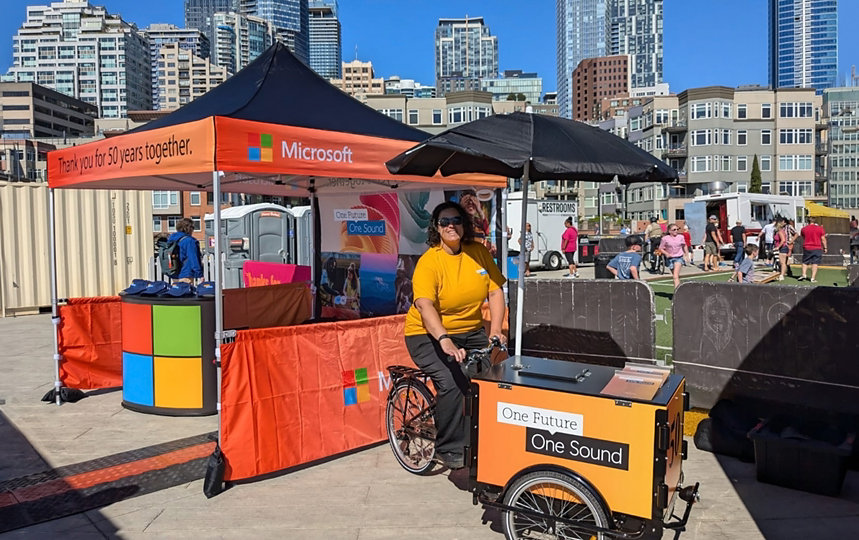 A woman with a tricycle cart at an outdoor event, with city buildings in the background.