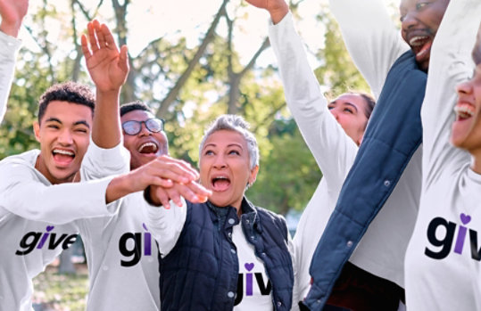 People outdoors in white “give” t-shirts celebrating with their hands in the air.