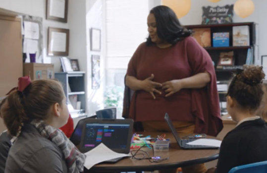 En lærer henvender sig til studerende, der sidder ved et bord med laptops foran sig.
