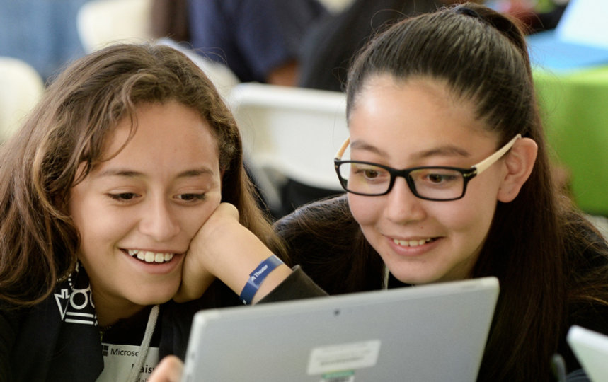 Two young girls sit together and play on a device.