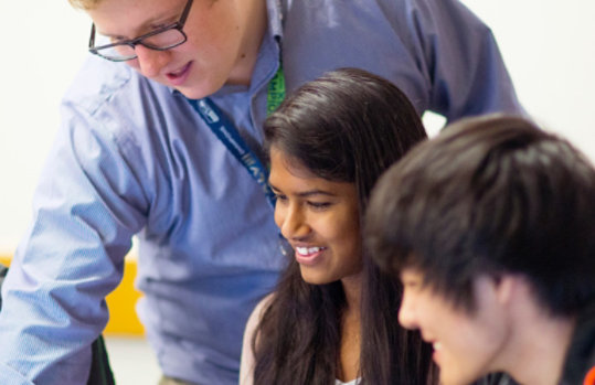 A group of students look at a monitor while a teacher points at the screen.