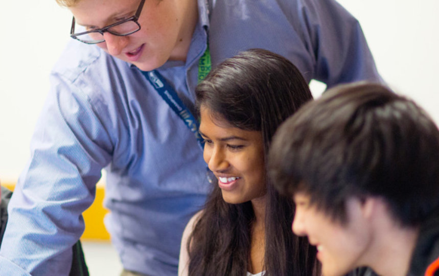 A group of students look at a monitor while a teacher points at the screen.