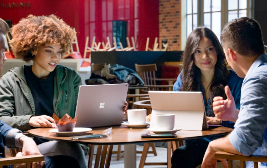 A group of young adults sit around a table talking to each other while working on devices.