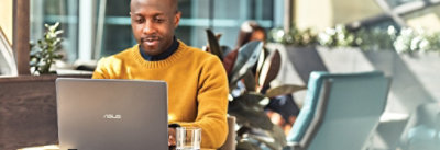 Muškarac u žutom džemperu sedi za stolom na kojem se nalazi čaša vode i radi na ASUS laptopu u moderno opremljenom prostoru.