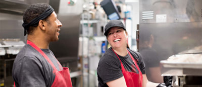 שני אנשים חובשים כובעים שחורים, סינרים רדומים וחולצות אפורות, מחייכים ומנהלים שיחה במטבח רועש עם מדפים