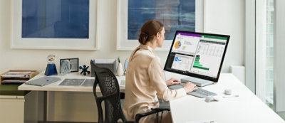 Osoba sedi za stolom u kancelariji i radi na računaru, a na velikom monitoru je otvoreno nekoliko dokumenata.