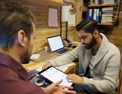 Dvojica muškaraca rade za stolom, pri čemu jedan drži tablet, a drugi nešto pokazuje. Laptop, knjige