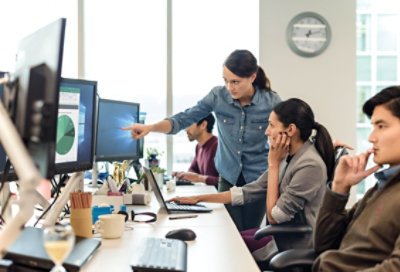  people working at a shared desk in an open office space. One person is standing and pointing at a desktop display monitor while a seated person observes.