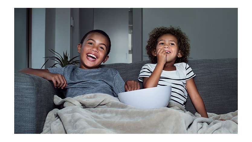 Duas crianças dividem uma tigela de salgadinhos enquanto assistem a um filme debaixo dos cobertores em casa