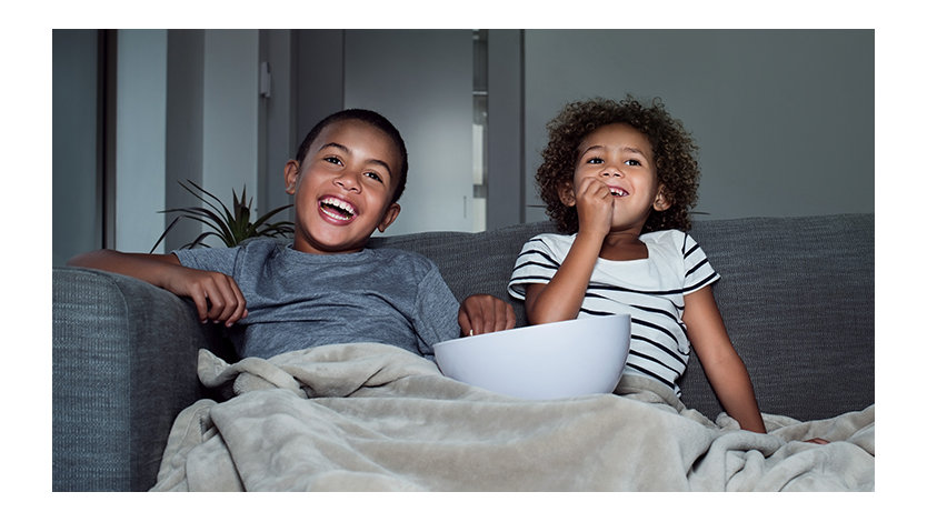Two children share a bowl of snacks and a blanket while enjoying a movie at home