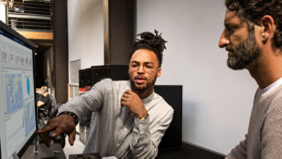 Două persoane uitându-se la un monitor desktop mare și discutând.