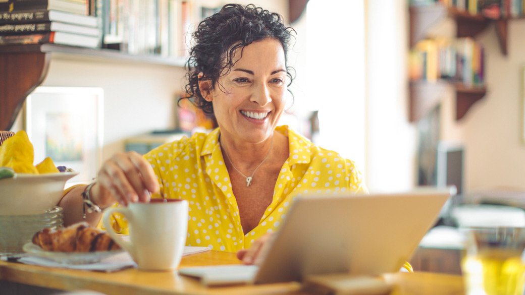 Smiling woman using laptop while having breakfast at her kitchen table.
