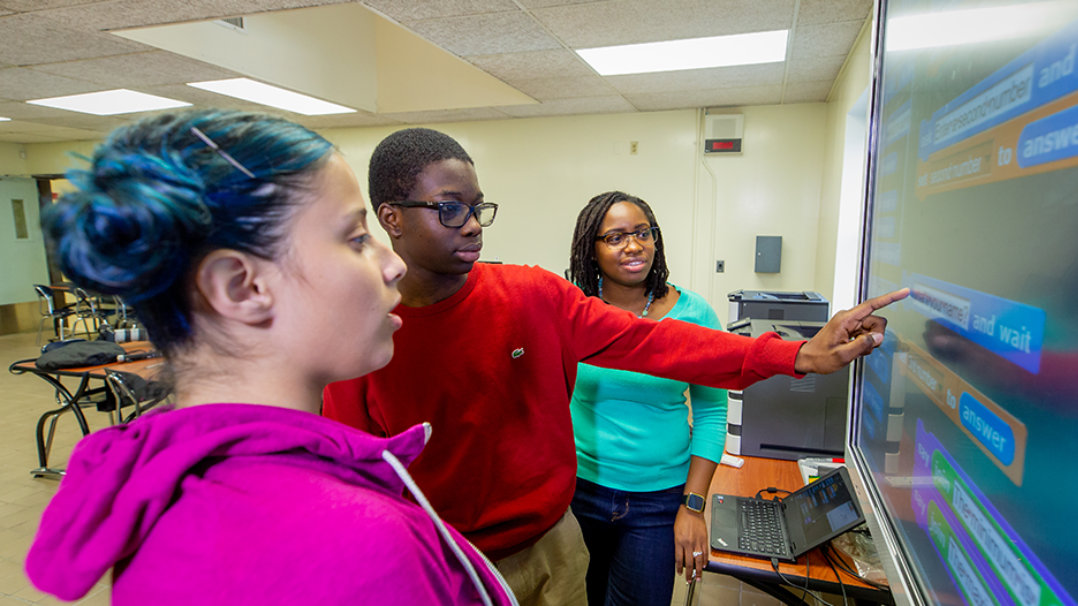 Two CS students collaborating near a large screen with a TEALS teacher’s assistant nearby.