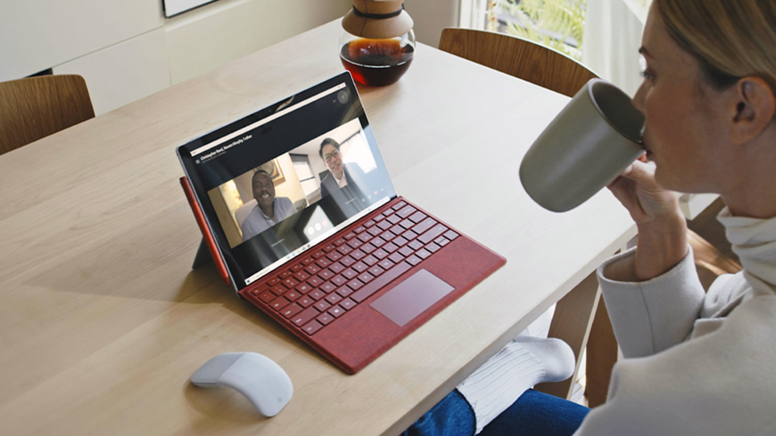 Woman using a Surface Pro 7 with a poppy red type cover, dialed into a teams call and drinking coffee