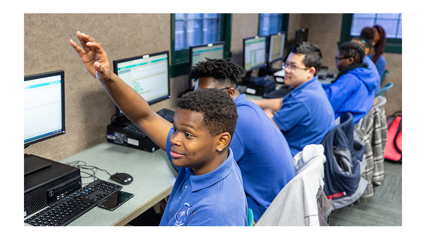 Student with hand raised in a classroom, with other students in front of computers.