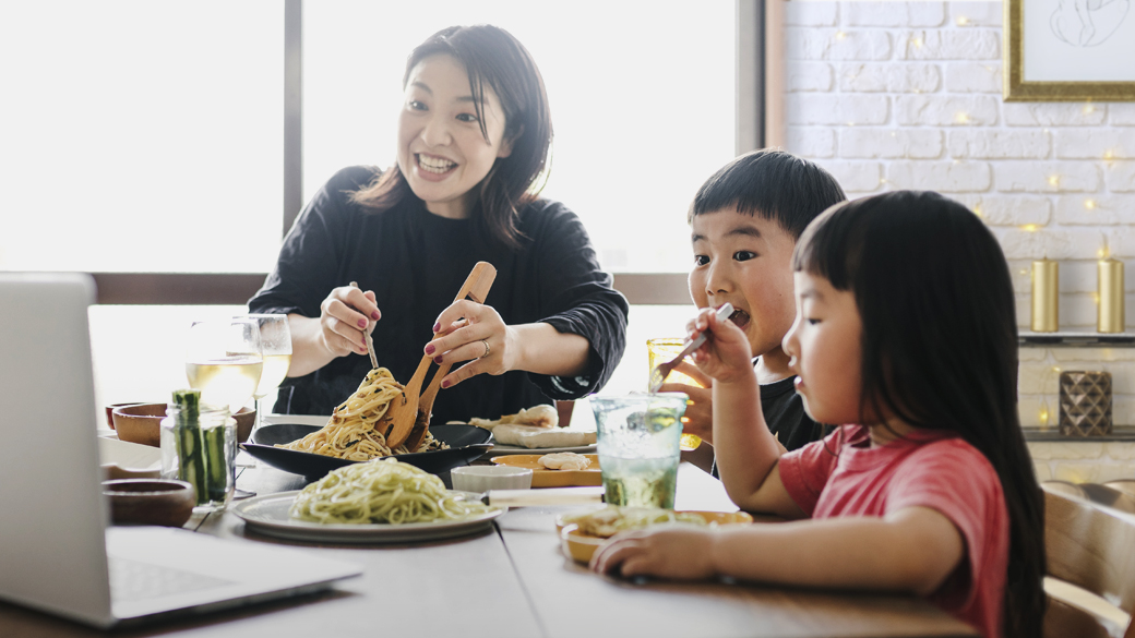 Mother and two children eat a meal together in kitchen while conducting video call on laptop computer