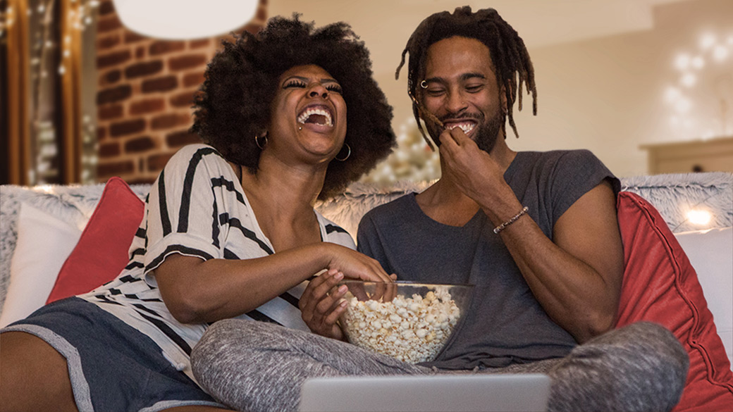 Laughing man and woman eat popcorn together on the couch at home while watching a video conference on laptop computer